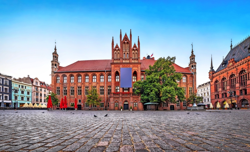 Historisches Backsteingebäude mit Türmen auf einem gepflasterten Platz.