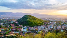 Blick auf Stadt mit bewaldetem Hügel und Bergen im Hintergrund bei Sonnenuntergang.