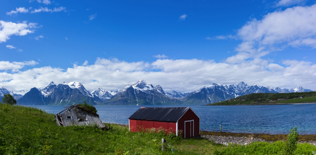 Rotes Haus vor Fjord und schneebedeckten Bergen, bewölkter Himmel.