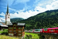 Glacier Express vor Alpenlandschaft mit Kirche und Holzhaus im Bündner Oberland.