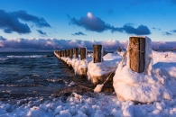Verschneite Holzpfähle ragen ins Meer unter blauem Himmel.