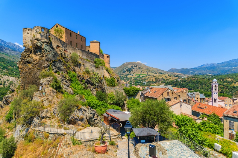 Burg auf Felsen über einem Dorf in einer bergigen Landschaft unter klarem Himmel.