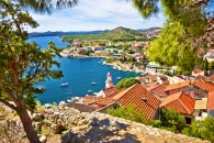 Küstenstadt mit roten Dächern und Leuchtturm am blauen Wasser, umgeben von Hügeln und Bäumen.