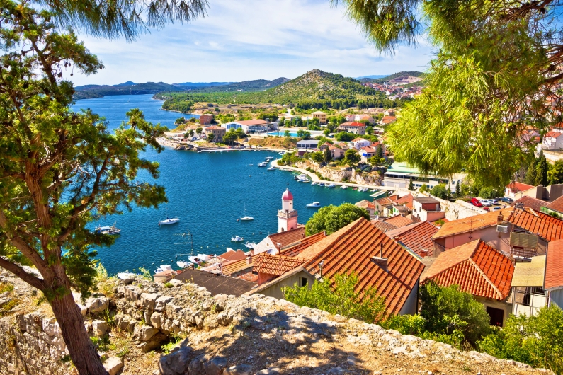 Küstenstadt mit roten Dächern und Leuchtturm am blauen Wasser, umgeben von Hügeln und Bäumen.
