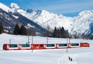 Ein roter Glacier Express Zug fährt durch eine schneebedeckte Landschaft mit Bergen im Hintergrund.