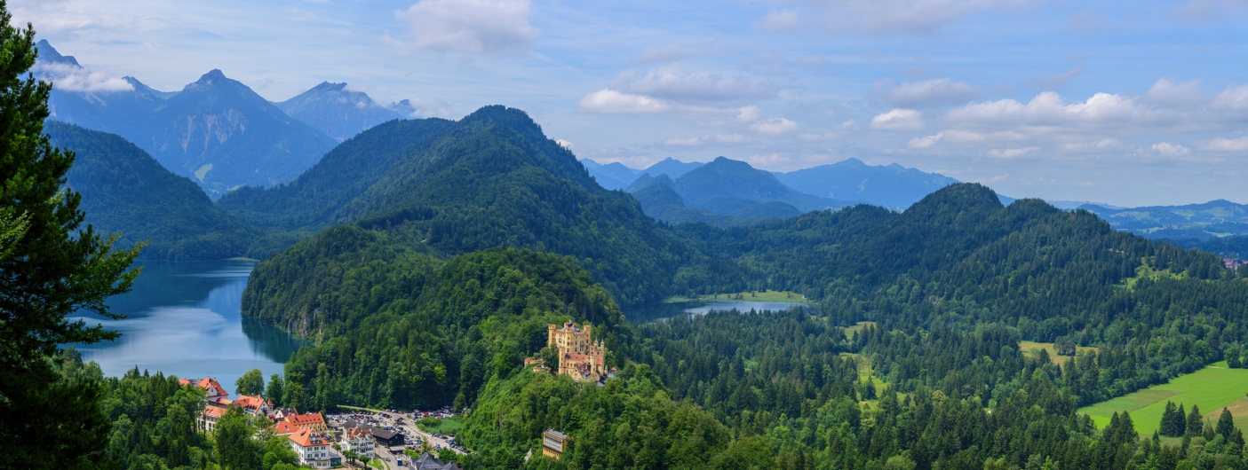 Schloss Hohenschwangau am Alpsee in Bayern, umgeben von bewaldeten Hügeln und Bergen.