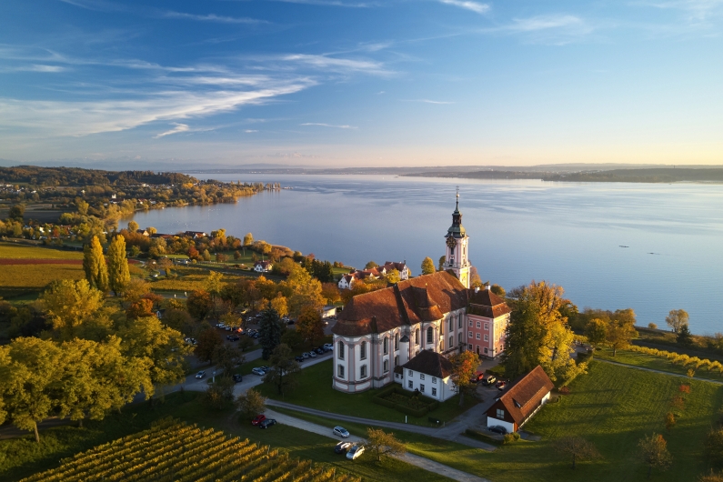 Kirche am Bodensee bei Sonnenuntergang, umgeben von Bäumen und Gewässer.