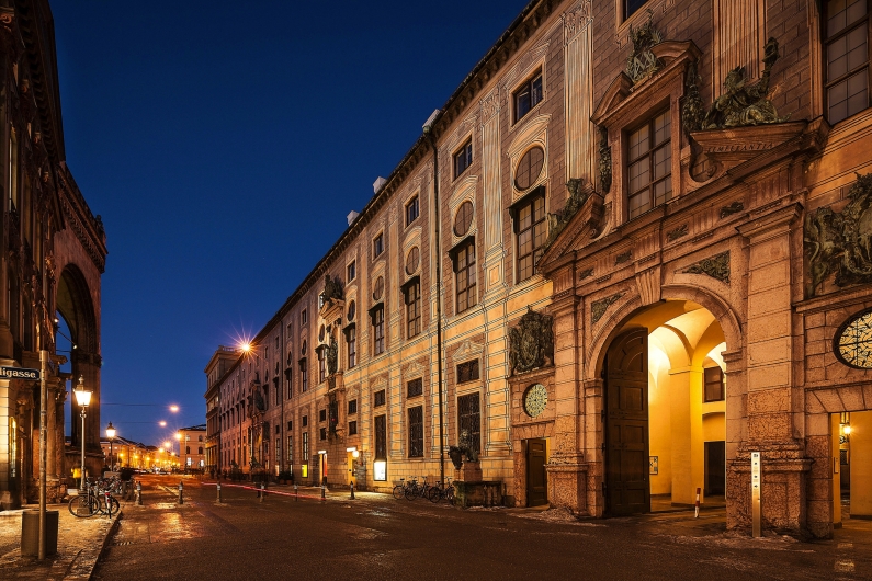 Fassade der Münchner Residenz bei Nacht, beleuchtet, mit Arkaden und dekorativen Elementen.