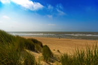 Strand mit Meer, Sanddünen und Gras unter blauem Himmel.