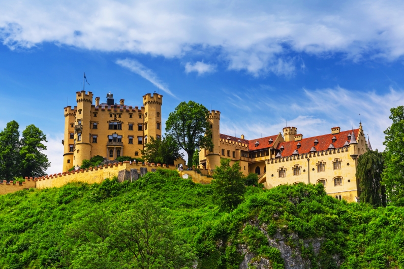 Schloss Hohenschwangau in Bayern, umgeben von grüner Landschaft und blauem Himmel.