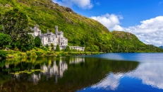 Schloss am See mit bewaldetem Hügel im Hintergrund, blauer Himmel mit Wolken.