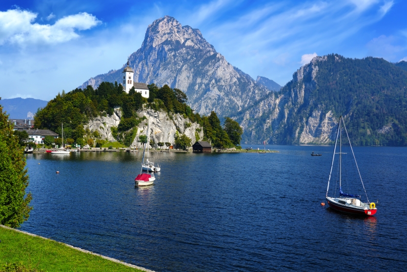 Boote auf einem See vor einer Kirche auf einem Hügel, im Hintergrund ein Berg.