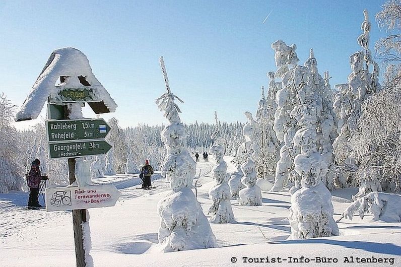 Verschneite Winterlandschaft mit Wegweiser und Spaziergängern im Hintergrund.