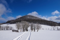 Verschneite Landschaft mit Hügel, Wald und blauem Himmel im Hintergrund.