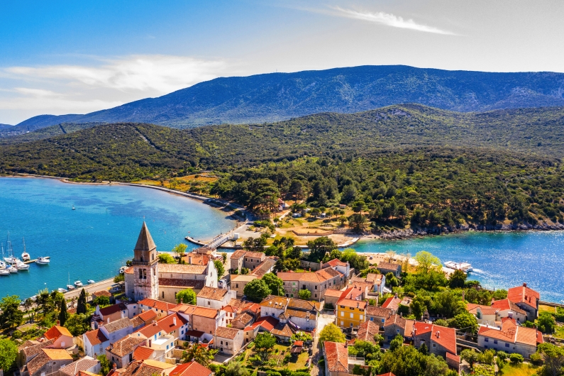 Luftaufnahme einer Küstenstadt mit Kirche, Meer und Bergen im Hintergrund.