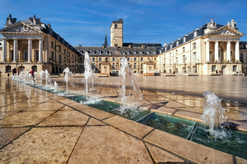 Platz mit Brunnen vor historischem Gebäude bei sonnigem Wetter.