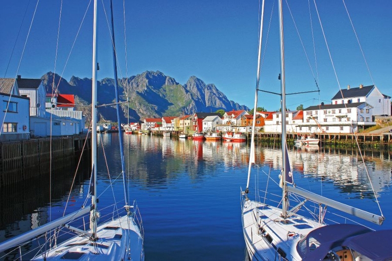 Segelboote in einem Hafen mit malerischen Häusern und Bergen im Hintergrund.