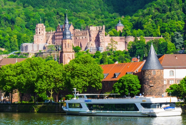 Schiff auf Fluss vor Heidelberger Schloss und grüner Hügellandschaft.
