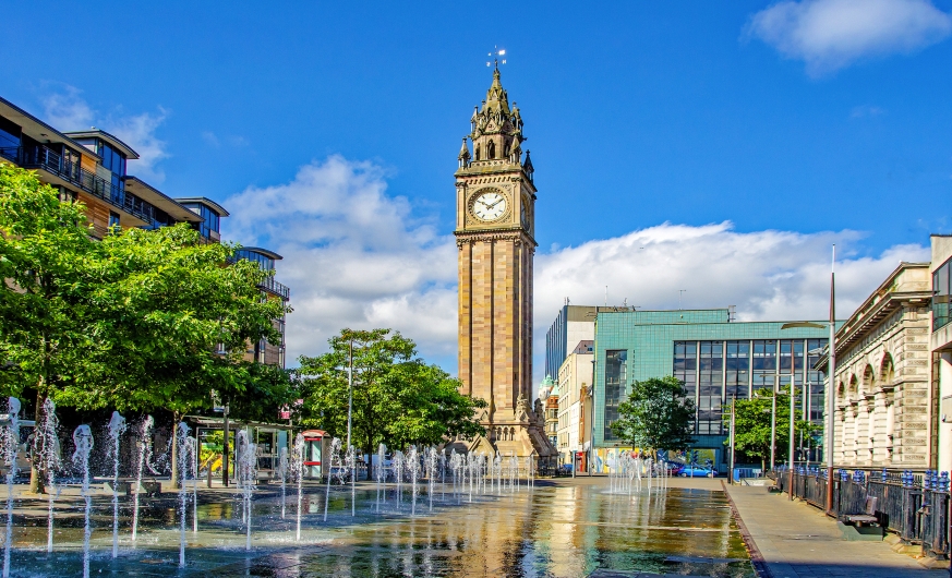 Historischer Uhrturm mit Wasserspielen und Gebäuden im Hintergrund, bei sonnigem Wetter.