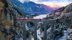 Roter Zug auf einem Viadukt in verschneiter Berglandschaft bei Sonnenuntergang.