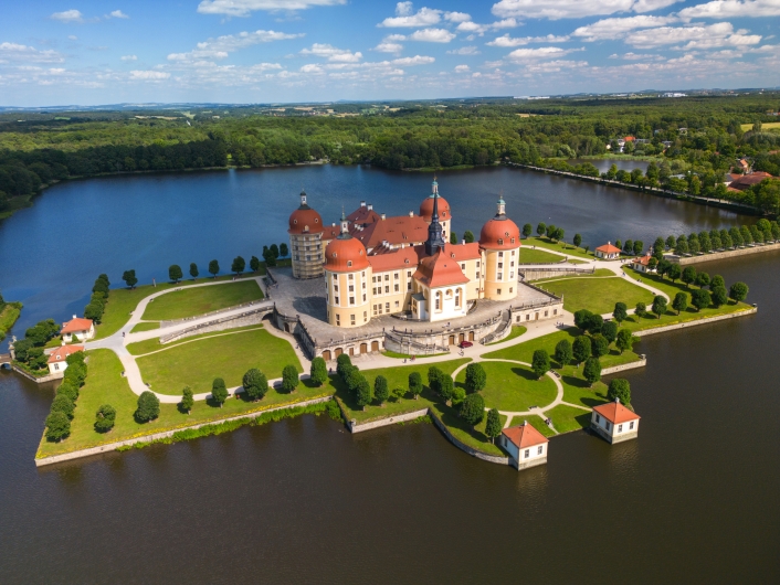 Luftaufnahme von Schloss Moritzburg in Sachsen, umgeben von einem Teich und grüner Landschaft unter blauem Himmel im Frühling.