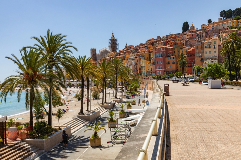 Uferpromenade mit Palmen vor bunten Häusern und einem Strand bei sonnigem Wetter.