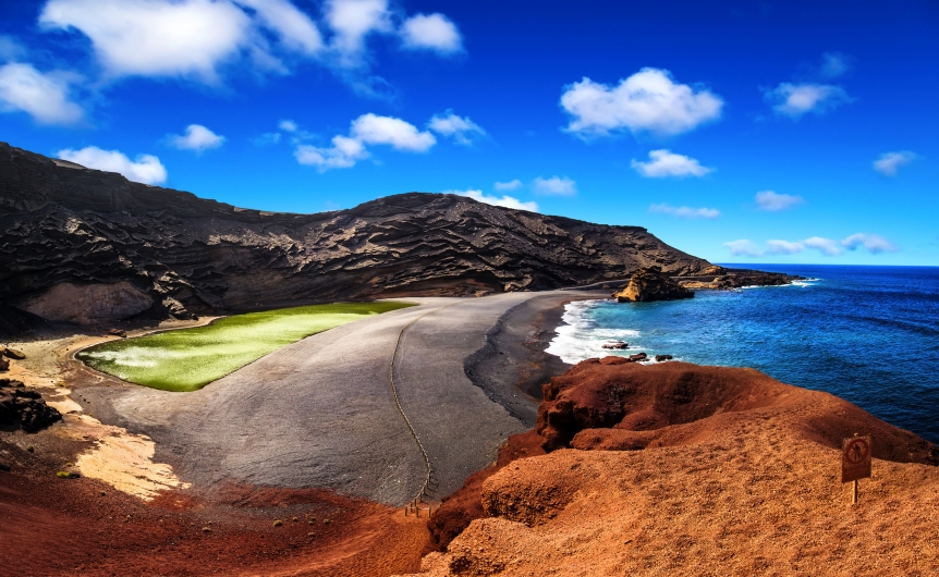 Küstenlandschaft mit grünem See, schwarzem Strand und blauem Himmel.
