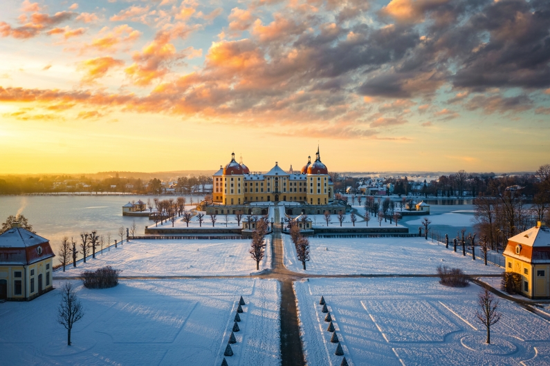 Schloss im Winter bei Sonnenuntergang, umgeben von verschneiten Gärten und einem See.