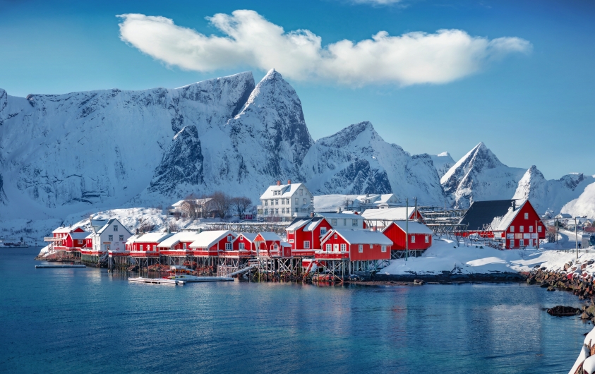 Verschneite Holzhäuser am Fjord vor schneebedeckten Bergen in Norwegen.