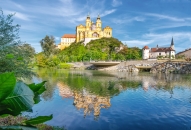 Barockes Schloss auf Hügel spiegelt sich im ruhigen Fluss unter blauem Himmel.