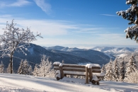 Verschneite Berglandschaft mit Bank im Vordergrund und blauen Himmel.