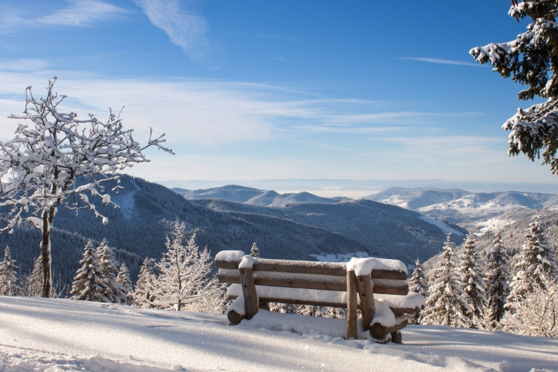 Verschneite Berglandschaft mit Bank im Vordergrund und blauen Himmel.