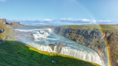 Wasserfall mit Regenbogen und grünes Umland bei sonnigem Himmel.