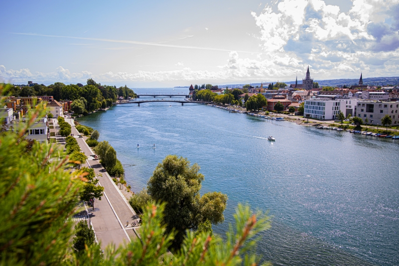 Stadt am Fluss mit Brücke, Gebäuden und Natur im Hintergrund bei sonnigem Wetter.