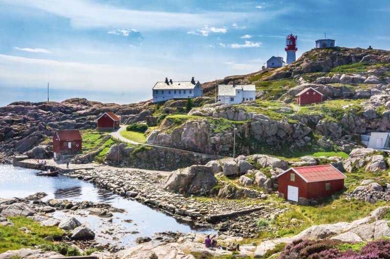 Küstenlandschaft mit Felsen, Häusern und Leuchtturm unter blauem Himmel.