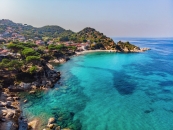 Küste mit blauem Meer und bewaldetem Hügel im Hintergrund.