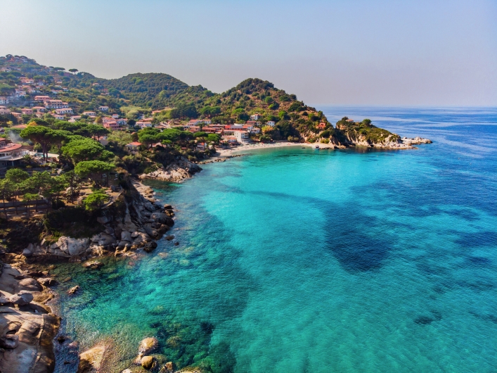 Küste mit blauem Meer und bewaldetem Hügel im Hintergrund.