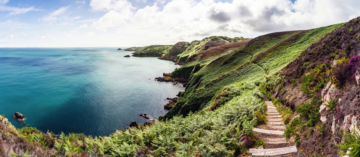 Küste mit grünem Hügel, Meer und Wolken im Hintergrund.