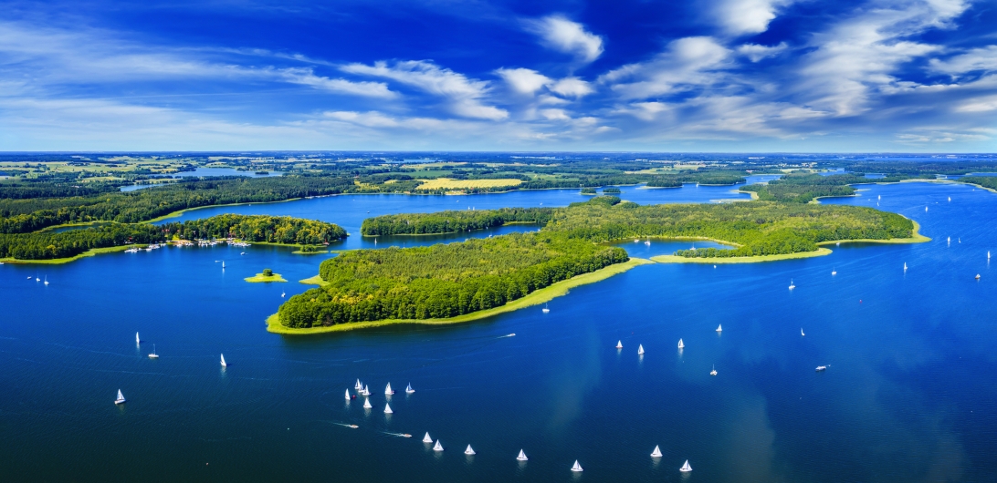 Luftaufnahme eines Sees mit Inseln und zahlreichen Segelbooten unter blauem Himmel.