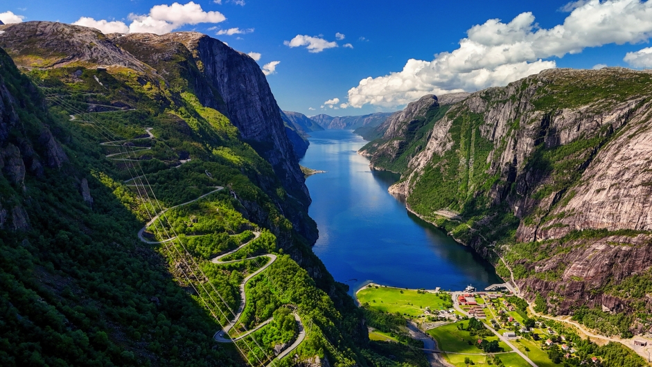 Blick auf einen Fjord mit steilen Bergen und einer kurvenreichen Straße bei sonnigem Wetter.