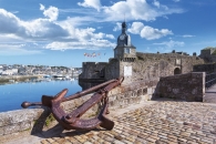 Historische Stadtmauer mit großem Anker, Hafen und Uhrturm im Hintergrund.