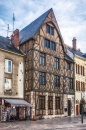 Fachwerkhaus in einer europäischen Stadt mit steilem Dach und mehreren Stockwerken, umgeben von anderen historischen Gebäuden. Im Erdgeschoss befinden sich Fenster und ein Zeitungskiosk.