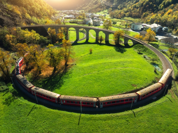 Luftaufnahme eines roten Zuges auf Kurve nahe einem Viadukt in grüner Landschaft.