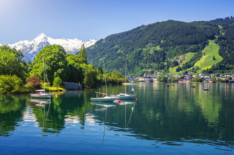 Bergsee mit Segelbooten, umgeben von Bergen und Wald, unter klarem Himmel.