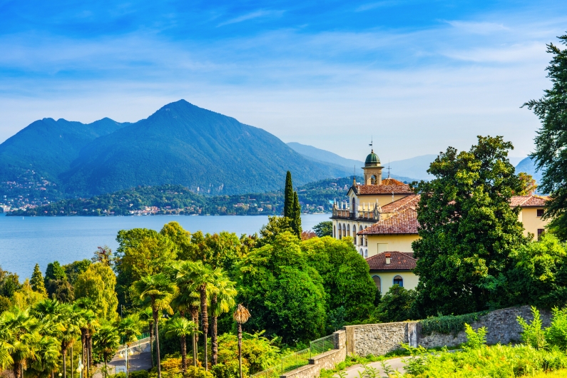 Landschaft mit Palmen, Villa und Blick auf einen See und Berge im Hintergrund.