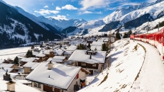 Schneesiedlung in Alpentälern mit vorbeifahrendem roten Zug und Bergen im Hintergrund.