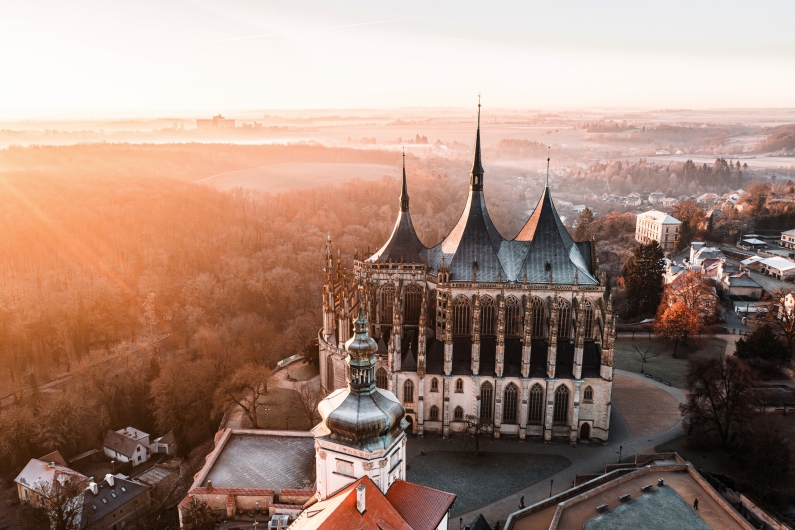 Luftaufnahme von einer gotischen Kathedrale, umgeben von herbstlichen Wäldern bei Sonnenaufgang.