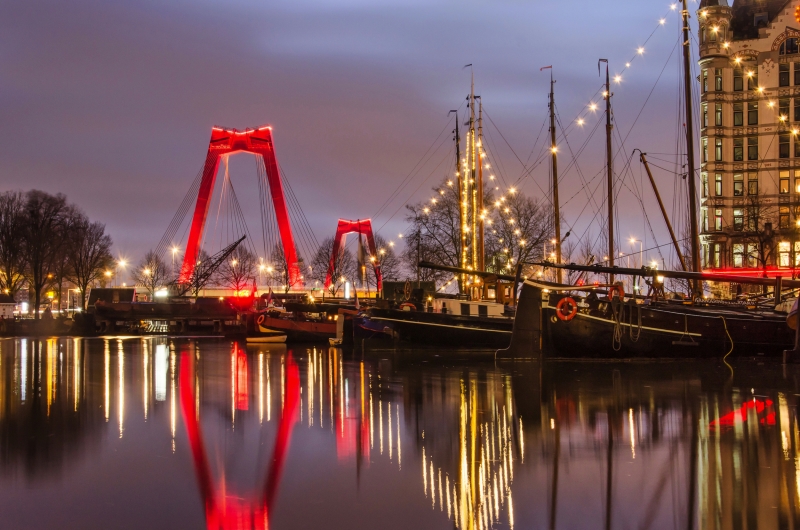 Abendliche Szenerie mit beleuchteter Brücke und Segelbooten im Hafen.