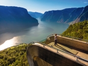 Person auf Aussichtsplattform mit Blick auf Fjord und Berge bei klarem Wetter.
