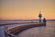 Eine weitläufige, schneebedeckte Mole führt in einem sanften Bogen zum grün-weißen Leuchtturm (Molenfeuer) von Travemünde. Direkt neben dem Turm steht ein geschmückter Weihnachtsbaum am Ende der Hafenmauer. Der Himmel ist in die warmen, orangefarbenen Töne eines winterlichen Sonnenuntergangs getaucht, während das ruhige Wasser der Ostsee den Kai umgibt.